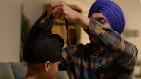 Father-Tying-Turban-Onto-Head-Of-Young-Sikh-Son-With-Top-Knot-Sitting-On-Sofa-At-Home-2